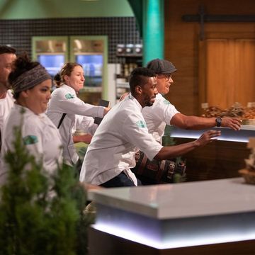 Two men and two women in white chef’s coats lunge and reach forward toward counter on Top Chef set.